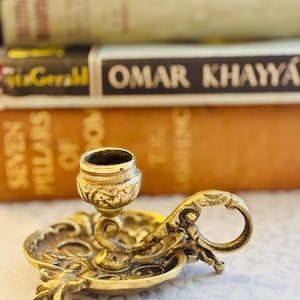 May include: A small, circular brass candle holder featuring an ornate, embossed floral pattern on the base and a curved handle. The central cup is designed to hold a single taper candle, with intricate metalwork detailing throughout the piece.