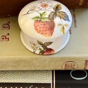 May include: A round white porcelain trinket box featuring a hand-painted botanical design of red strawberries, green leaves, and small white flowers on both the lid and the base. The ceramic container has a smooth, glossy finish and a domed lid.