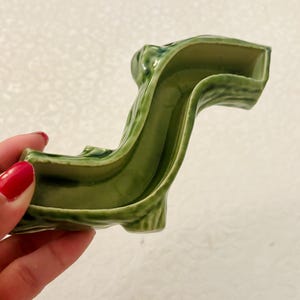 May include: A glossy, olive-green ceramic object with a curved, elongated shape and a hollow centre. The surface features a textured, organic finish with subtle variations in the green glaze, creating a smooth, reflective appearance throughout.