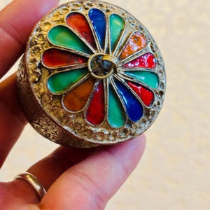 May include: A round, silver-toned metal container featuring an intricate, embossed floral pattern on the sides. The lid is decorated with a radial pattern of teardrop-shaped inlays in vibrant shades of red, orange, green, and blue.