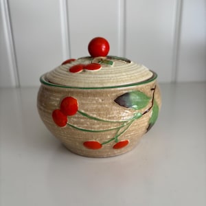 May include: A ceramic sugar bowl with a lid, featuring a beige textured finish and a cherry design. The lid is topped with a red cherry. Green leaves and red cherries are painted on the bowl's exterior. The bowl is on a white surface.