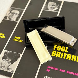 May include: A silver harmonica with a black plastic case and a small, rectangular, pale yellow cleaning cloth. The items are placed on a black and white album cover featuring the text FOOL BRITANNIA, written and devised by LESLIE BRICUSSE & ANTHONY NEWLEY.