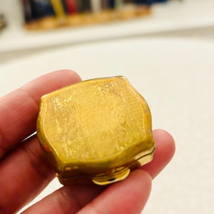 May include: A small, square-shaped gold-toned metal compact case with rounded corners and a textured, engraved floral pattern on the lid. The compact features a smooth, polished gold-coloured latch on the front edge for opening and closing.