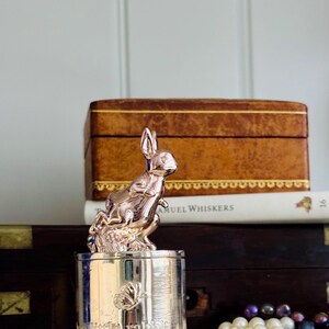 May include: A silver container with a rose gold bunny figurine. The container has the words "my little rabbit" and a butterfly design. The bunny is perched on top of the container. The container is on a white surface.