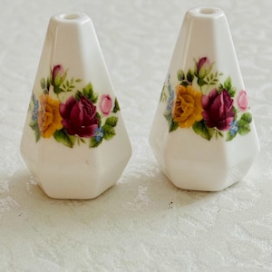 Könnte beinhalten: Ein Paar Salz- und Pfefferstreuer aus weißer Keramik mit facettierter, konischer Form. Jeder Streuer ist auf der Vorderseite mit einem Blumenmotiv verziert, das gelbe und dunkelrote Rosen, grüne Blätter sowie kleine blaue Blüten zeigt.