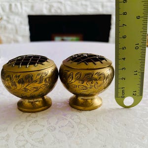 May include: Two round brass flower frogs featuring etched leaf patterns and a wire grid top. A yellow measuring tape is positioned next to the items, showing a height of approximately 6.5 cm for each brass vessel with a pedestal base.