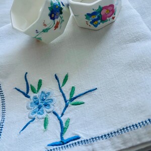 May include: Two white, octagonal ceramic napkin rings with floral designs in blue, pink, and orange. They rest on a white linen napkin with blue embroidered floral details and a blue border.