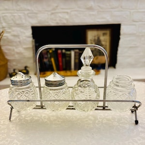 May include: A condiment set featuring four clear glass containers with a diamond-cut pattern held in a silver-toned metal wire caddy. The set includes two shakers with perforated metal lids, one bottle with a pointed glass stopper, and one open jar.