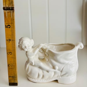 May include: A white ceramic planter shaped like a boot, with a sculpted figure of a child holding a sack. The planter is approximately 5 inches tall, with a detailed design and a glossy finish.