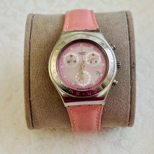 May include: A Swatch chronograph watch with a pink dial and a pink leather strap. The watch has a silver-colored case and features three sub-dials. The date is displayed at the 3 o'clock position. The watch is resting on a gray surface.