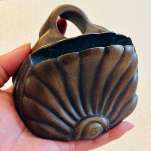 May include: A dark bronze-toned wall pocket or hanging vessel shaped like a scallop shell. The object features a rounded, fan-like design with radiating ridges and a circular opening at the top for mounting or hanging on a vertical surface.