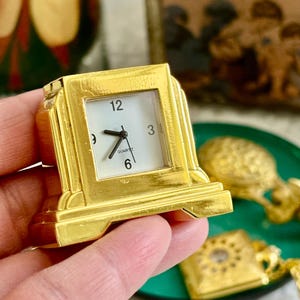 May include: A small gold-toned quartz clock with a square frame and a white face. The clock face features black numerals at the 12, 3, 6, and 9 positions, black hour and minute hands, and the word QUARTZ printed in black below the centre.
