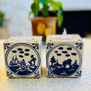 May include: Two square ceramic candle holders featuring a white base with hand-painted blue landscape designs inside circular borders. Each cube has a circular opening at the top designed to hold a small tea light candle, with floral corner accents.