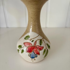 May include: A small ceramic vase with a beige speckled neck and a white body. The vase is decorated with a red and blue floral design and green leaves. The vase is approximately 13 cm tall.