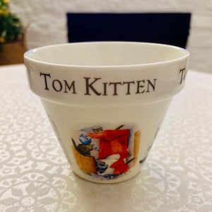 May include: A white ceramic planter with a flared rim featuring the text Tom Kitten in dark brown lettering. The front of the pot displays a colorful illustration of a cat in a blue hat and a red garment, set against a light background.