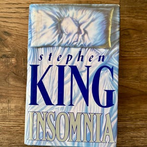 May include: A hardcover book titled "Insomnia" by Stephen King. The cover features a person's profile on a pillow, with blue and white wavy patterns. The author's name and title are in bold, blue and white lettering.