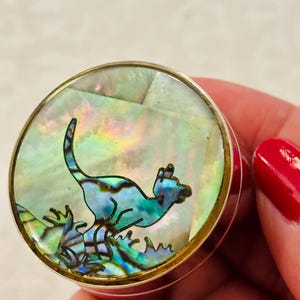 May include: A round metal pill box featuring an inlaid iridescent abalone shell top. The inlay displays a dark silhouette of a kangaroo standing on a textured ground against a shimmering, multi-coloured pearlescent background with green, pink, and blue tones.