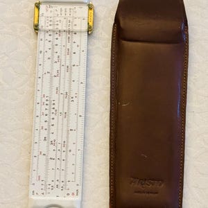 May include: A white Aristo slide rule featuring a clear plastic cursor, gold-tone metal fittings, and detailed numerical scales, alongside a dark brown leather protective case embossed with the Aristo brand name and Made in Germany.