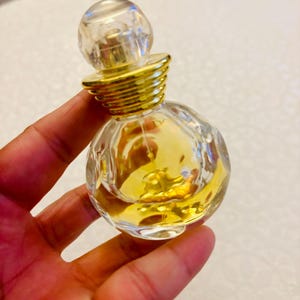 以下が含まれることがあります： 黄金色の液体が入った、小さく丸い透明なガラス製の香水瓶。瓶にはファセットカットが施された球状のガラス製ストッパーと、リブ状の金色の金属製カラーが付いています。ガラス製の本体は、多面的なテクスチャーデザインが特徴です。