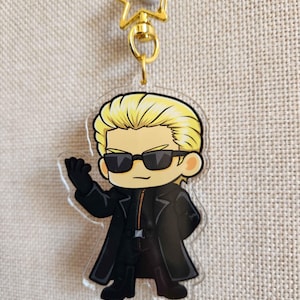 Op de afbeelding: Een heldere acryl sleutelhanger met een stripfiguur met blond haar, een zonnebril en een zwarte outfit. De sleutelhanger heeft een gouden stervormige bovenkant en een gouden ring om sleutels aan te bevestigen.