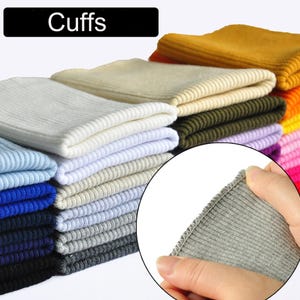 Op de afbeelding: Een stapel geribbelde gebreide manchetten in verschillende kleuren, waaronder wit, blauw, grijs en geel. Het woord "Cuffs" wordt weergegeven in een zwarte rechthoek. Een close-up toont de rand van de grijze manchet.