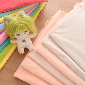 Può includere: Una bambola di peluche con capelli verdi e orecchie da gatto, con un fiocco, accanto a una pila di tessuto dai colori pastello. Il tessuto comprende sfumature di rosa, pesca e crema, con un arcobaleno di altri colori visibili.