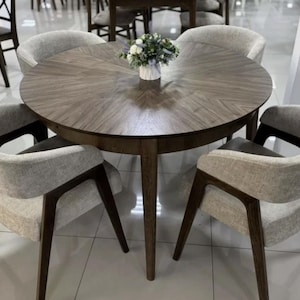 May include: A round wooden dining table with a dark brown finish and a chevron wood grain pattern, accompanied by four upholstered chairs featuring light gray fabric seats and dark brown wooden frames, with a small floral arrangement on top.