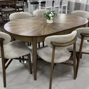 Puede incluir: Una mesa de comedor ovalada de madera con un patrón de chapa en forma de rayos de sol en la superficie, acompañada de seis sillas con marcos de madera oscura, asientos tapizados en beige claro y respaldos curvos, con un arreglo floral central.