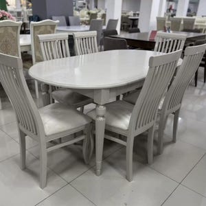 Peut inclure: Une table à manger ovale avec une surface gris clair lisse et quatre pieds cannelés, accompagnée de six chaises assorties. Chaque chaise présente un dossier à lattes verticales et un coussin d'assise gris clair, formant un ensemble cohérent.