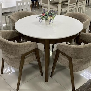 May include: A round dining table with a white top and dark wood legs, surrounded by four upholstered chairs with curved backs and dark wood frames. A small vase with artificial flowers sits in the center of the table surface.