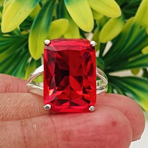 May include: A silver ring featuring a large, rectangular, faceted red gemstone held in a four-prong setting. The band is split into two thin, polished silver arms that meet at the base of the stone, creating a clean and symmetrical design.