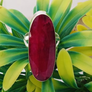 May include: A ring featuring a large, oval-shaped, deep red gemstone with a faceted surface. The stone is set in a simple metal band that encircles the base of the gem, providing a secure hold for the elongated, vibrant red centerpiece.