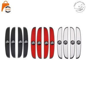 Puede incluir: Un conjunto de doce protectores de borde de puerta de coche dispuestos en tres grupos de cuatro, en colores negro, rojo y blanco. Cada protector es una tira larga y curva con un emblema circular del logotipo de Lexus en negro y plata.