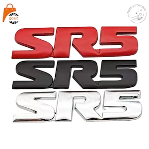 May include: Three automotive emblem badges featuring the text SR5 in a bold, stylized font. The top emblem is solid red, the middle emblem is matte black, and the bottom emblem has a reflective, polished chrome finish with a smooth surface.
