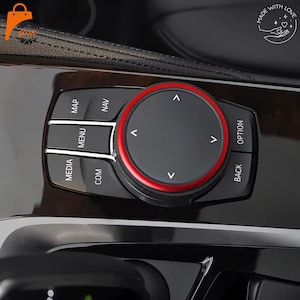 Può includere: Una manopola di controllo multimediale per auto nera con bordo circolare rosso e frecce direzionali. Il pannello della console include pulsanti etichettati MAP, NAV, MENU, MEDIA, COM, OPTION e BACK su una superficie nera lucida.