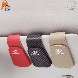 Puede incluir: Tres soportes para gafas de sol para visera de coche en rojo, patrón de fibra de carbono negro y beige. Cada soporte presenta el logotipo de Lexus en texto blanco y un mecanismo de cierre magnético sobre una visera gris claro.