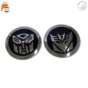 Puede incluir: Dos emblemas circulares con diseños plateados metálicos en relieve sobre un fondo negro brillante. El emblema izquierdo muestra la insignia Autobot, mientras que el derecho presenta la insignia Decepticon, ambos con bordes cromados.