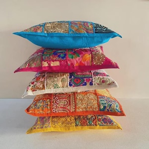 May include: A stack of five square throw pillows featuring patchwork designs with intricate embroidery, beadwork, and metallic thread accents. The pillows display a vibrant color palette including bright blue, magenta, cream, orange, and yellow.