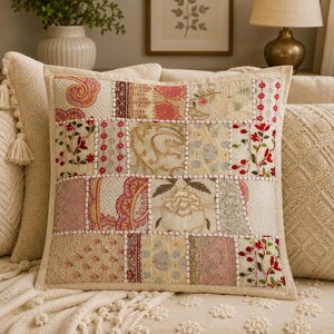 May include: A square throw cushion featuring a patchwork design of various embroidered fabric panels in shades of cream, pink, red, and green. The panels are joined by visible white stitching, showcasing floral, paisley, and geometric patterns.