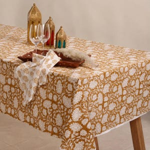 May include: A rectangular tablecloth featuring a mustard yellow background with a repeating white floral pattern. On top sits a dark brown wooden tray, two clear glass wine glasses, three gold-toned metal lanterns, and a white cloth napkin.