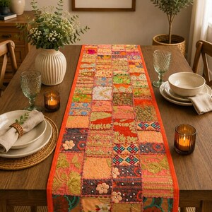 May include: A rectangular table runner featuring a patchwork design of various fabric squares in shades of orange, pink, red, and gold. The runner is decorated with intricate embroidery, small floral appliqués, and scattered white beads.