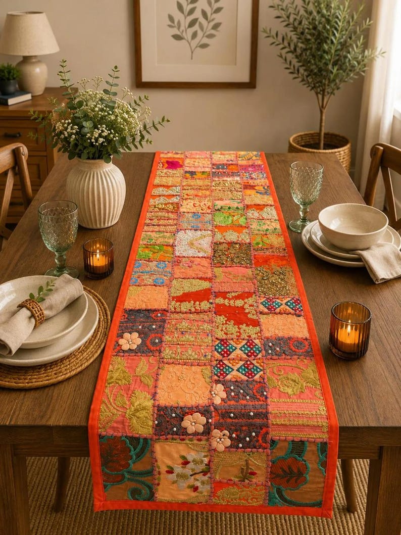 Op de afbeelding: Een rechthoekige tafelloper met een patchworkontwerp van diverse stoffen vierkanten in tinten oranje, roze, rood en goud. De loper is versierd met ingewikkeld borduurwerk, kleine bloemapplicaties en verspreide witte kralen.