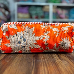 May include: A rectangular quilted pouch in a vibrant orange hue featuring a detailed floral and botanical print in beige and dark blue. The bag includes a zipper closure with a red and white striped trim along the top edge and a matching side strap.