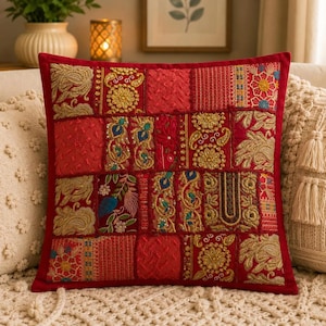 Könnte beinhalten: Ein quadratisches Zierkissen mit einem Patchwork-Design aus roten Stoffbahnen. Jedes Feld ist mit aufwendigen goldenen, blauen und grünen Stickereien verziert, darunter Blumen-, Paisley- und geometrische Muster, eingefasst von einem roten Rand.