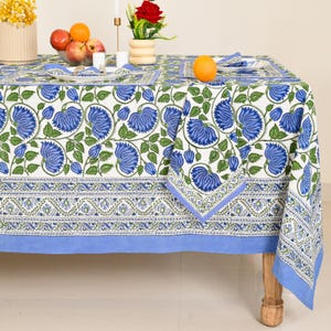May include: A white tablecloth and matching napkins featuring a repeating pattern of blue flowers and green leaves. The fabric has a wide border with a smaller floral vine design and a solid light blue hem. The set is displayed on a wooden table.