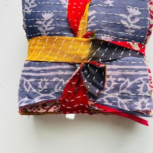 May include: A folded stack of fabric featuring a blue base with white floral patterns and white running stitch details. The stack is secured with a strip of red and yellow fabric tied around the center, creating a bundle of layered textiles.