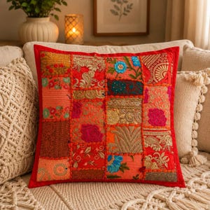 Puede incluir: Un cojín cuadrado con un diseño de patchwork compuesto por varios paneles de tela en tonos rojos, naranjas y dorados. La superficie está decorada con bordados intrincados, motivos florales y detalles en hilo dorado metálico, bordeado en rojo.