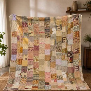 Op de afbeelding: Een grote rechthoekige quilt met een patchworkontwerp bestaande uit talloze kleine stoffen vierkantjes in diverse gedempte tinten zoals beige, geel, roze, bruin en zacht paars, allemaal aan elkaar genaaid met zichtbare stikpatronen.