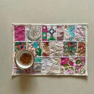May include: A rectangular placemat featuring a patchwork of embroidered fabric squares in various colours including pink, green, blue, and gold. A small white bowl with a gold rim containing brown almonds sits on the left side of the placemat.