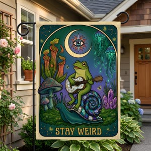 May include: A rectangular garden flag featuring a green frog playing a guitar while sitting on a large snail. The scene includes a crescent moon, an eye, jellyfish, pitcher plants, mushrooms, and purple crystals. The text STAY WEIRD is at the bottom.